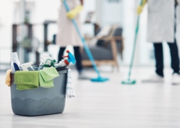 Eimer mit Reinigungsmitteln und Mikrofasertüchern im Vordergrund; im Hintergrund wischen zwei Reinigungskräfte den Boden in einem hellen Gebäude.