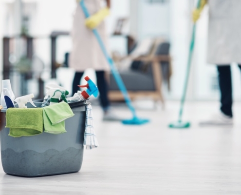 Eimer mit Reinigungsmitteln und Mikrofasertüchern im Vordergrund; im Hintergrund wischen zwei Reinigungskräfte den Boden in einem hellen Gebäude.