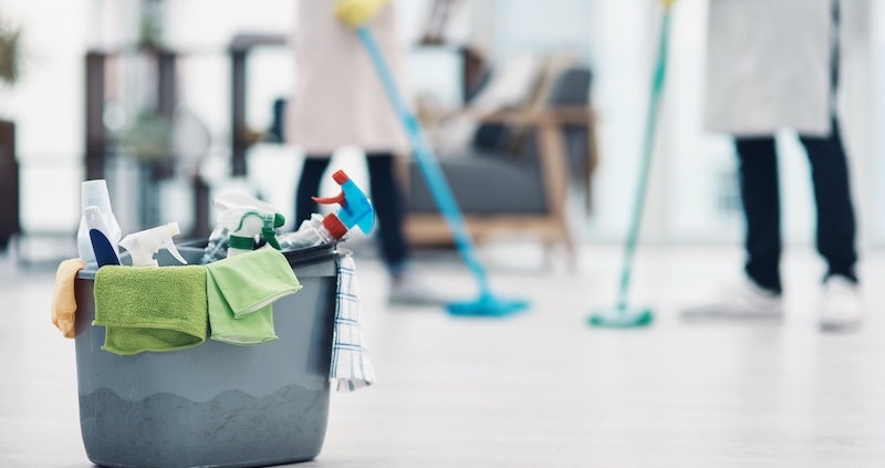 Eimer mit Reinigungsmitteln und Mikrofasertüchern im Vordergrund; im Hintergrund wischen zwei Reinigungskräfte den Boden in einem hellen Gebäude.