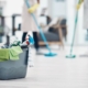 Eimer mit Reinigungsmitteln und Mikrofasertüchern im Vordergrund; im Hintergrund wischen zwei Reinigungskräfte den Boden in einem hellen Gebäude.